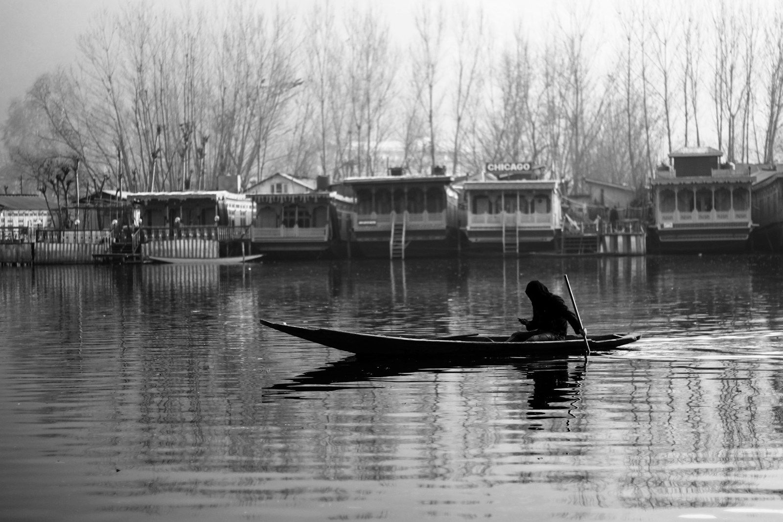 Black and white photo of a boatman paddling a shikara past houseboats on Dal Lake.